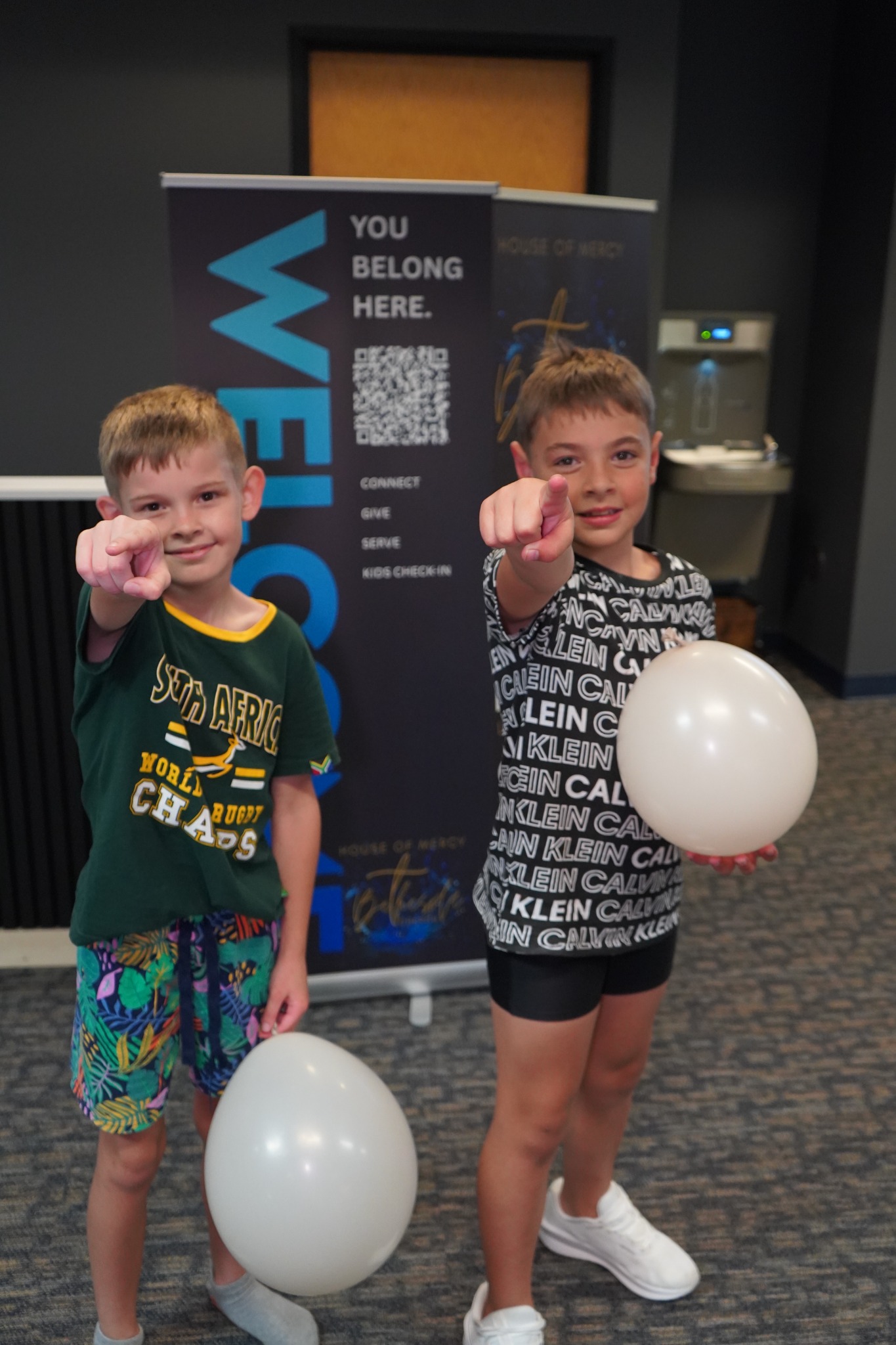 Two smiling children with balloons beside a church welcome sign.
